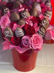 A charming arrangement of a dozen pink and red roses in a rustic bucket, accompanied by a dozen chocolate-covered strawberries in a decorative box.